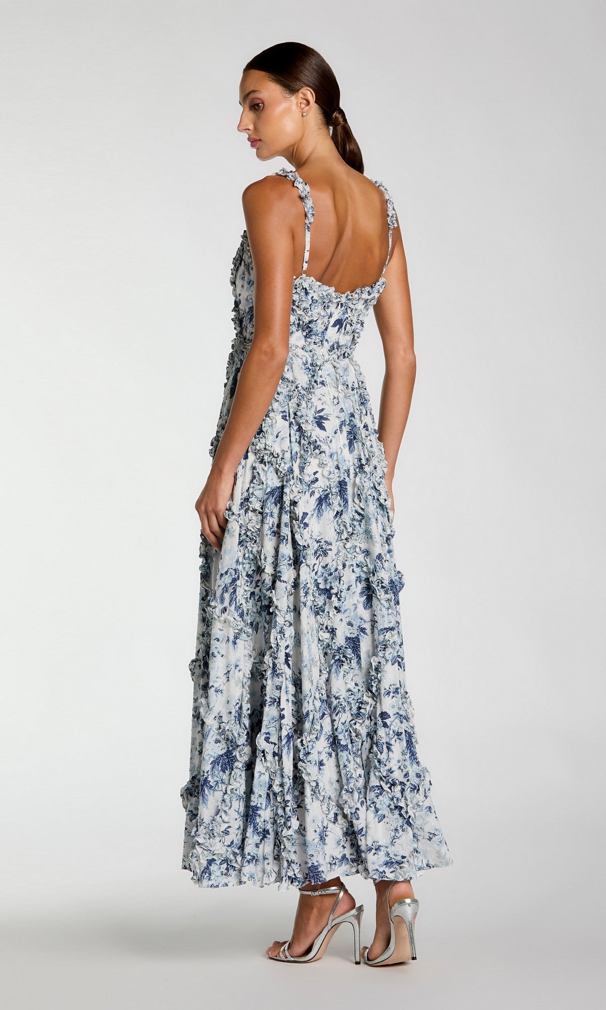 A woman faces away, wearing the Mac Duggal 8256 Floral Wedding Guest Dress with ruffled straps and hem in blue and white, paired with silver high-heeled sandals against a plain background.